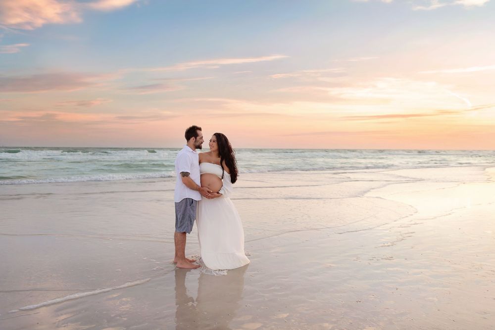Couple embracing on a sandy beach at sunset, woman in white dress, waves and colorful sky in background.