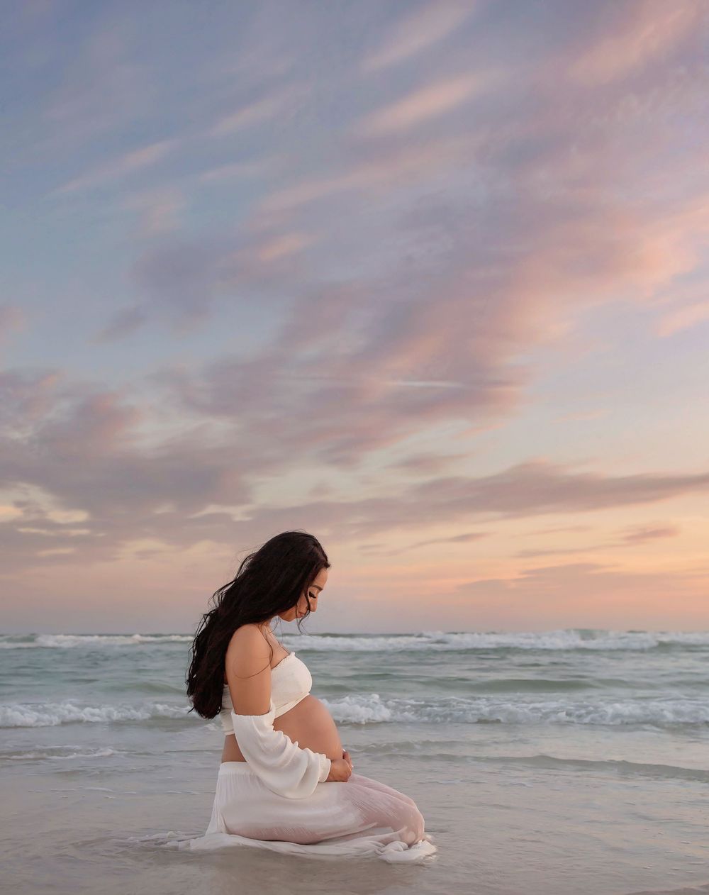 Pregnant woman in white dress kneeling on beach at sunset with pink clouds and ocean waves.