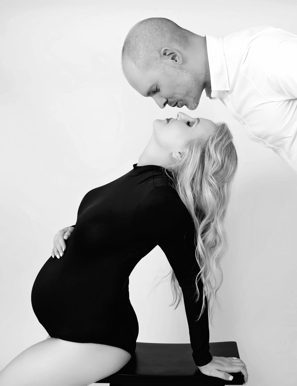 Black and white maternity photo of pregnant woman in black dress kissing partner in white shirt in studio.