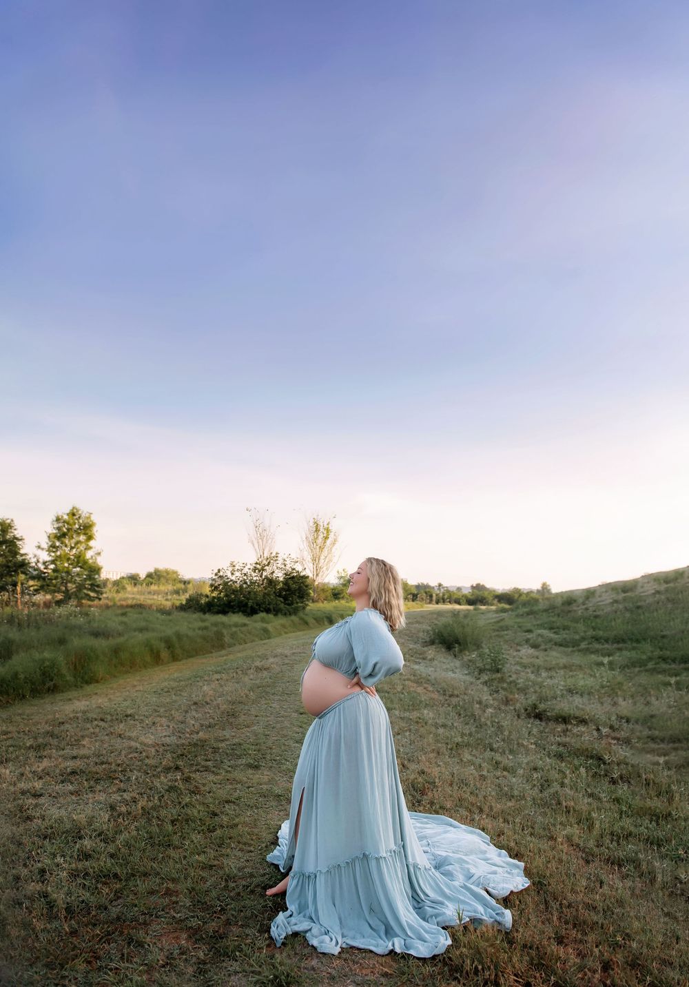 Maternity session in flowing blue dress on grassy hillside during golden hour.