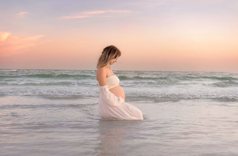 Woman in flowing white dress walking through ocean waves at sunset.