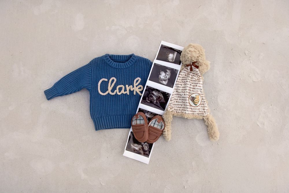 Blue knit sweater with name Clark, ultrasound photos, tiny shoes, and a pom-pom hat on a neutral background.
