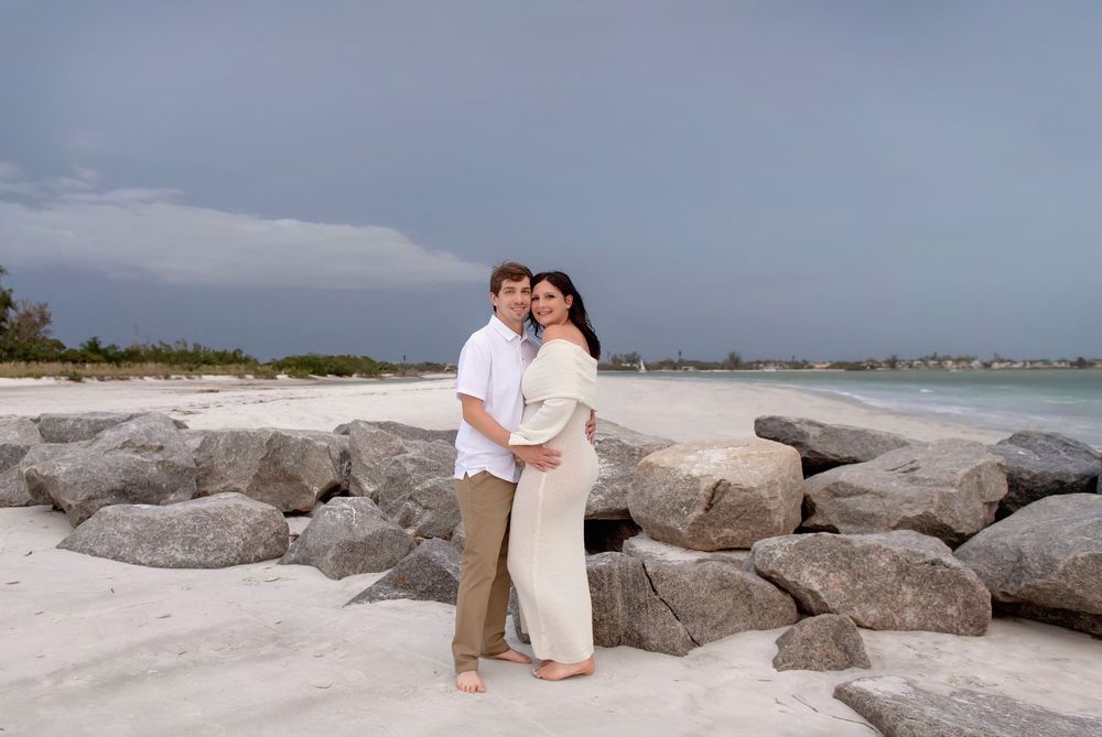 Couple embracing on a white sand beach with large rocks and cloudy sky backdrop.