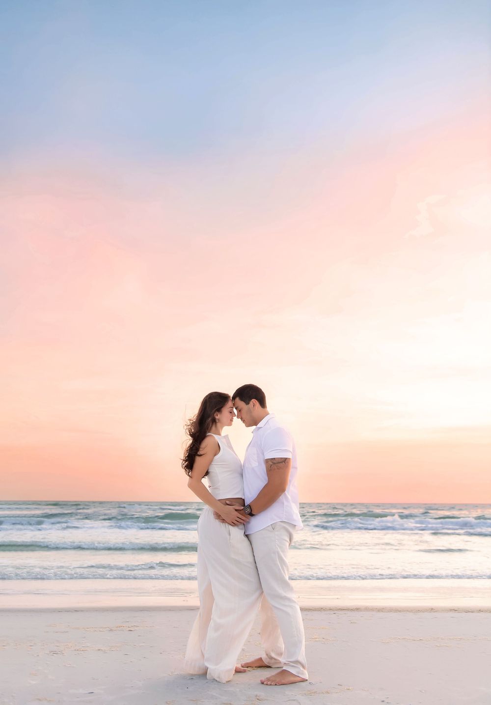 Couple in white embracing on a sandy beach at sunset with pastel pink and blue sky backdrop.