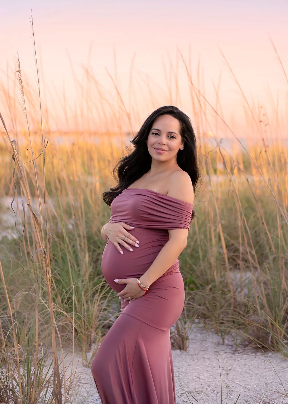 Pregnant woman in mauve off-shoulder gown poses on sandy beach dunes at golden sunset.