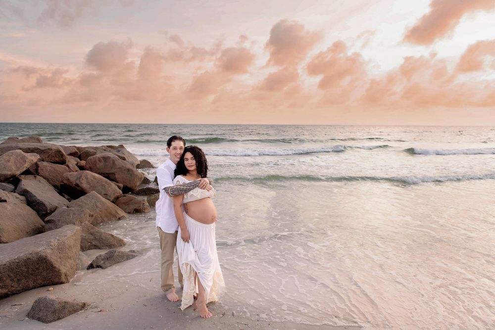 Couple poses on beach at sunset, man embracing pregnant woman in white dress near rocks and ocean waves.
