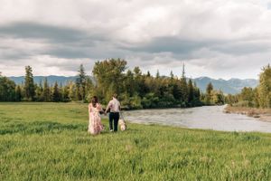 A couple walks through a lush green meadow with mountains and a river in the background.