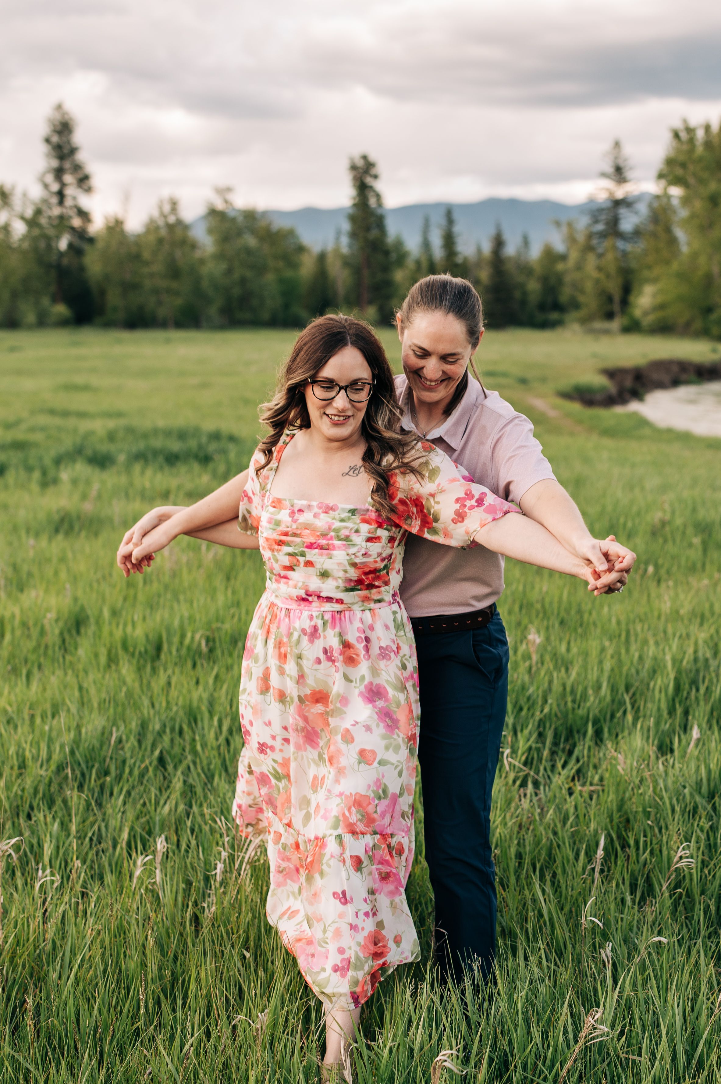 Two people share a joyful dance in a meadow with mountains and evergreens in the background.