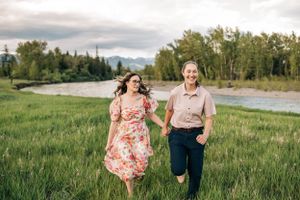 Two people run joyfully through a grassy field with mountains and trees in the background.