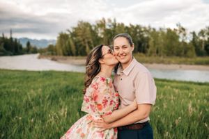 A romantic couple embraces in a grassy field with a river and mountains in the background during golden hour.