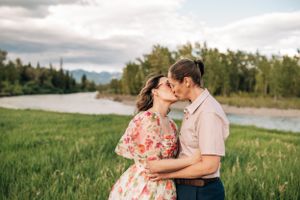 A loving couple shares a kiss in a meadow with a winding river and forest in the background.