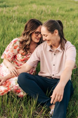 A couple shares a joyful moment while sitting in tall grass during an outdoor photoshoot.