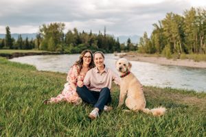 A couple sits with their golden retriever on grassy riverbank with trees and mountains in background.