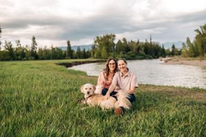A couple and their golden retriever relax on grass near a winding river with mountains in view.