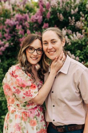 Two people pose together in front of flowering lilac bushes on a sunny day.