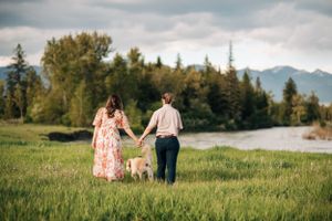 Two people hold hands while walking through a grassy field with mountains and river in background.