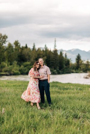 A couple stands together in a scenic mountain meadow beside a flowing river.