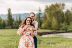 A couple embraces while standing in a green field with mountains and river in the distance.