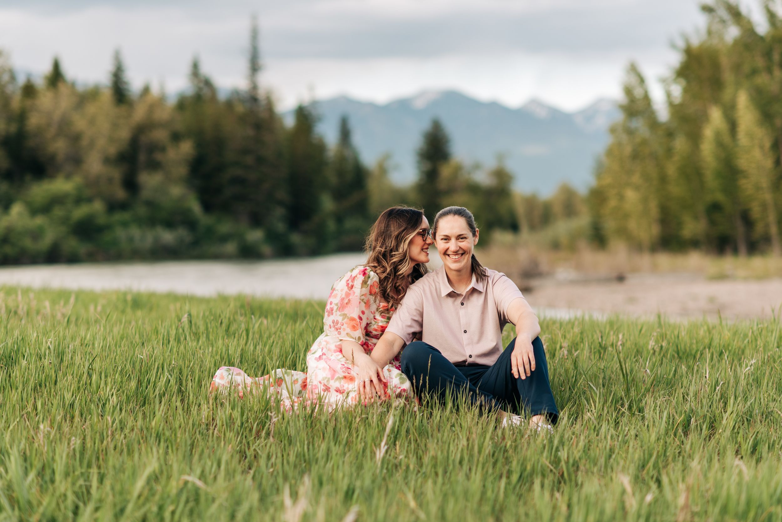 Two people sit together in tall grass with mountain peaks and river visible in background.