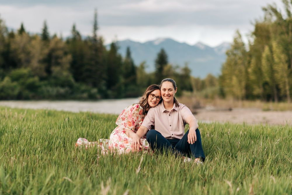 A couple sits together in a grassy meadow with mountains and evergreen trees in the background.