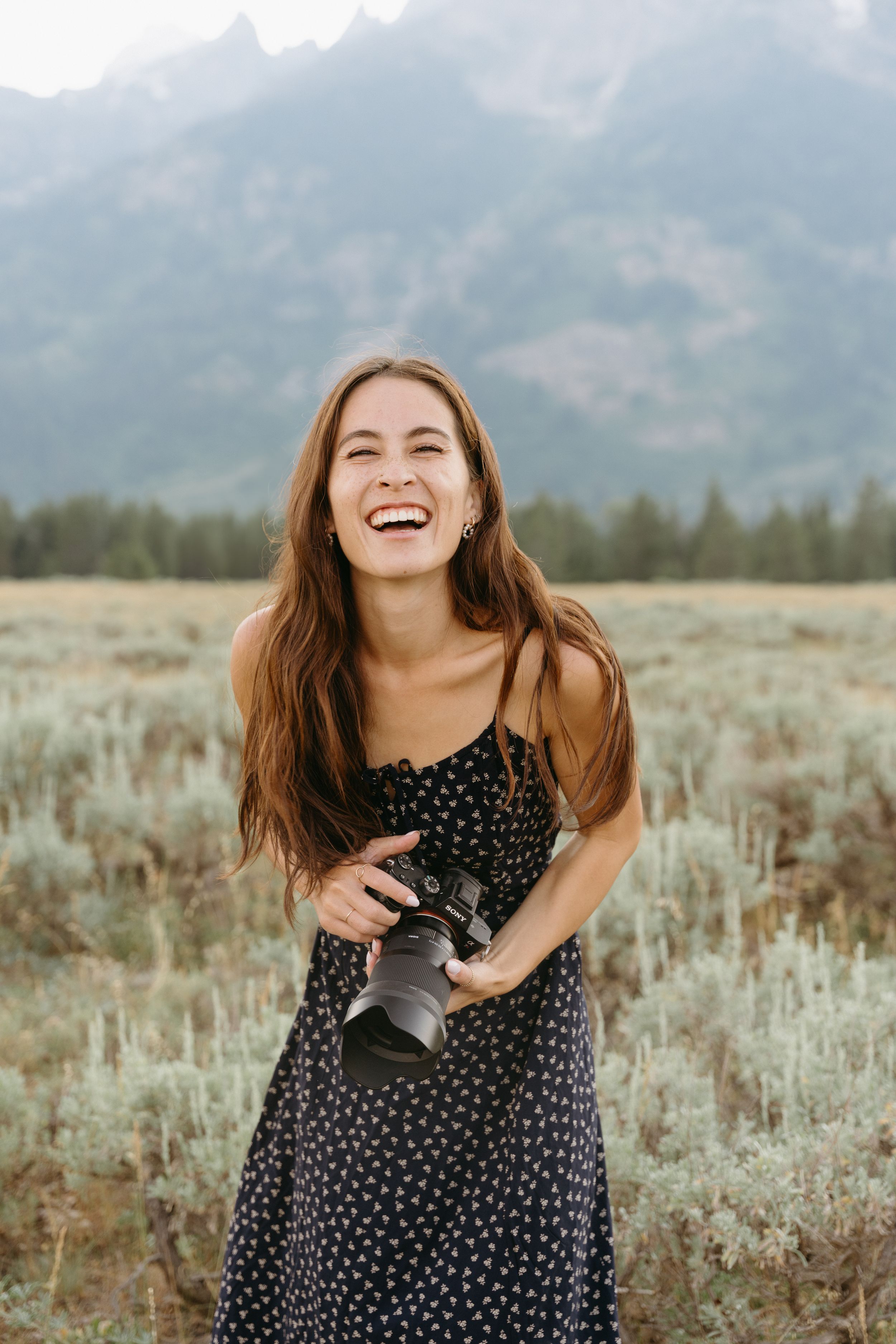Joyful person in polka dot dress laughing while holding camera in field with mountain backdrop.