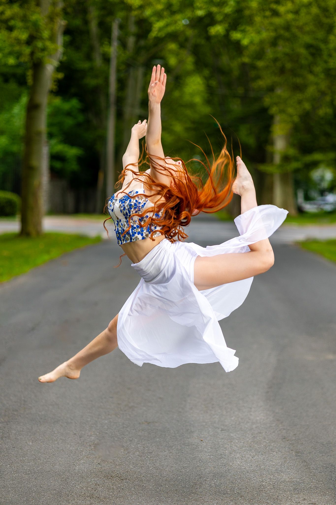 Dancer in white dress performing graceful mid-air leap on a tree-lined road.