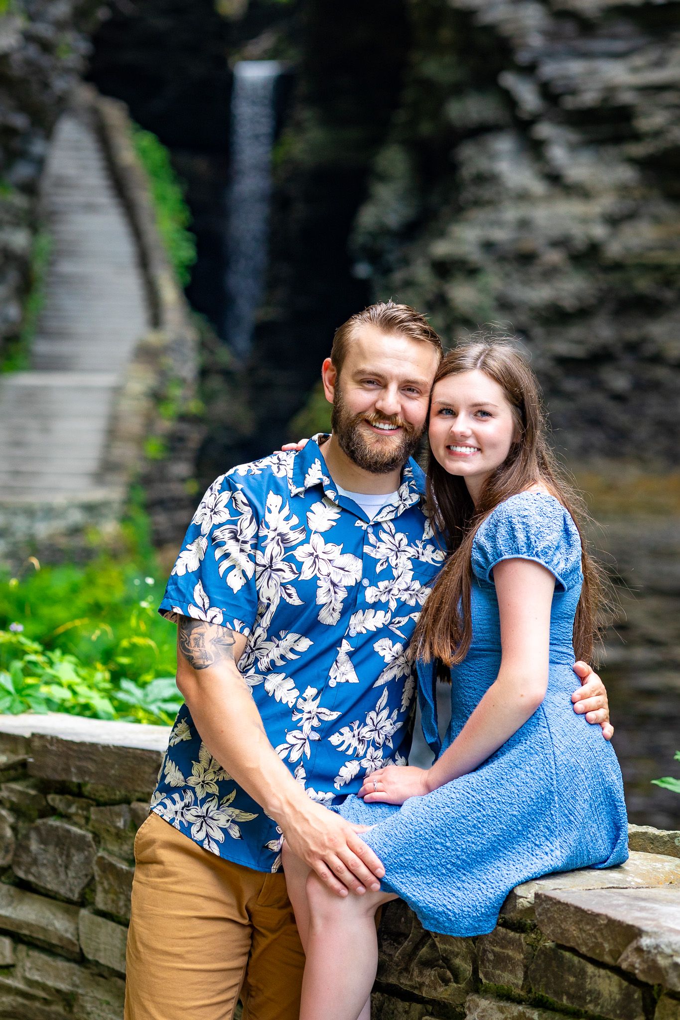 Couple in coordinating blue outfits pose together by stone wall near waterfall.