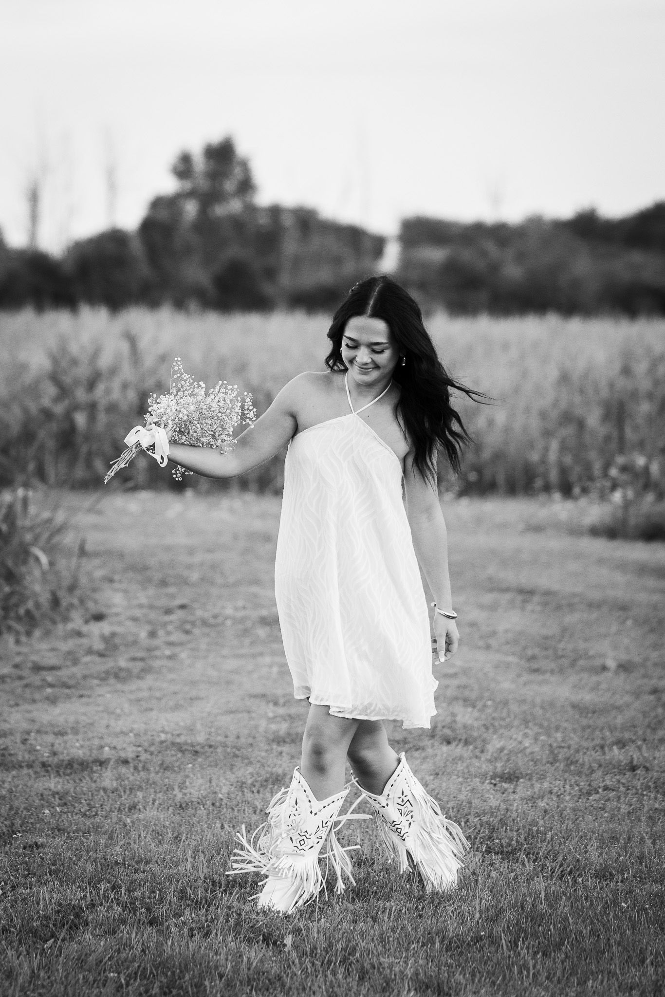 A person in a flowing white dress and fringe boots dances joyfully in a field during a black and white photoshoot.