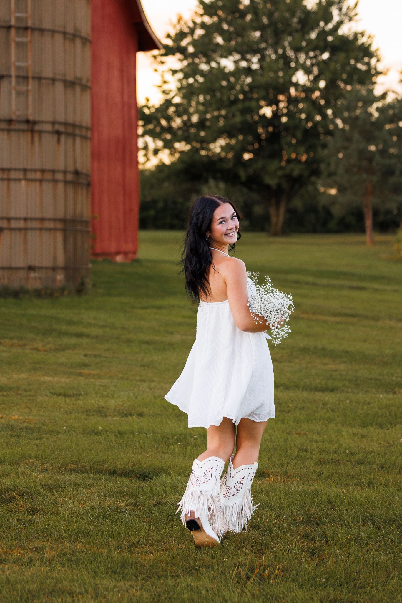 A woman in a white off-shoulder dress dances in a grassy field at sunset holding baby's breath flowers.