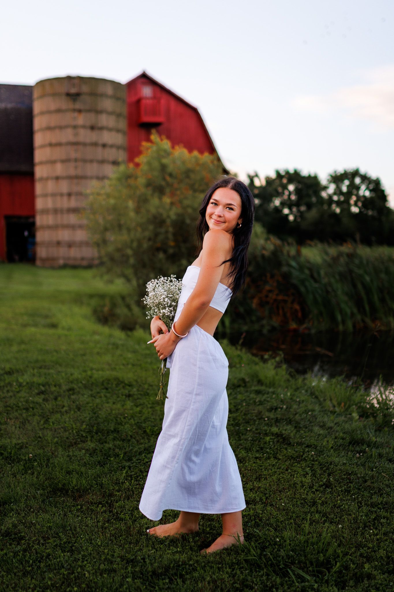 A person in a white dress twirls joyfully in a grassy field at sunset with a red barn and silo in the background.