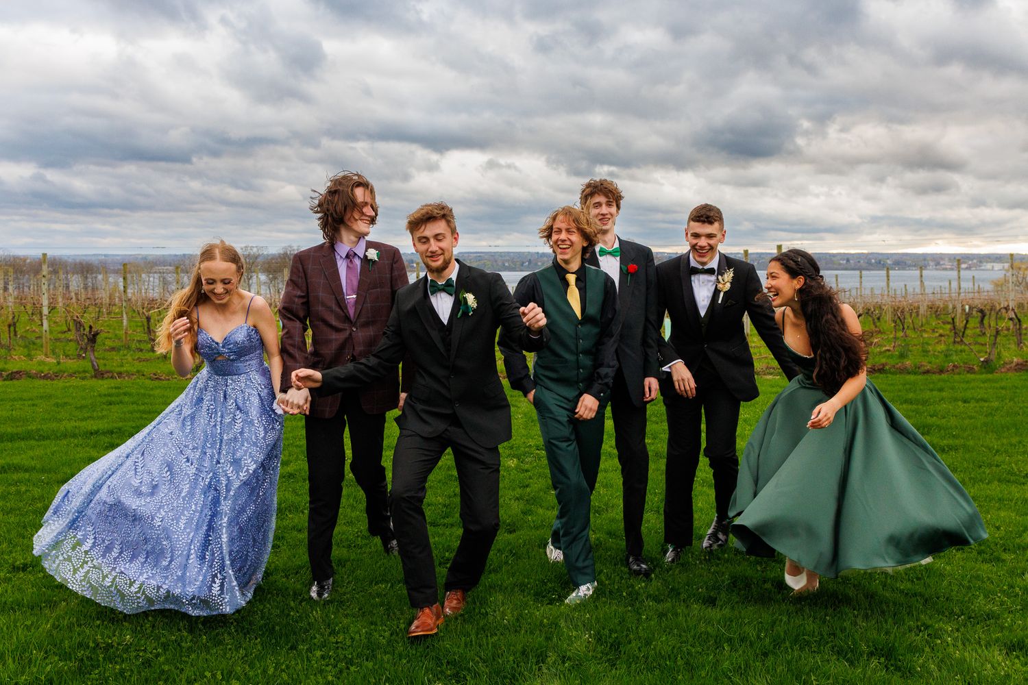 Wedding party in formal attire poses playfully in a green field under dramatic cloudy skies.