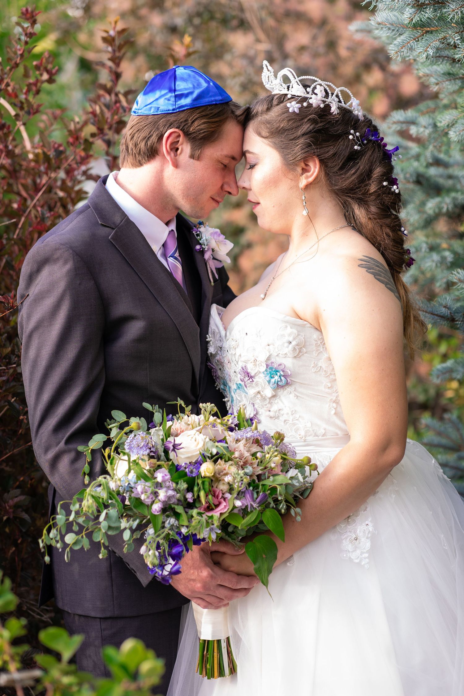 Groom wearing yarmulke and bride wearing tiara in an intimate pose with bouquet.