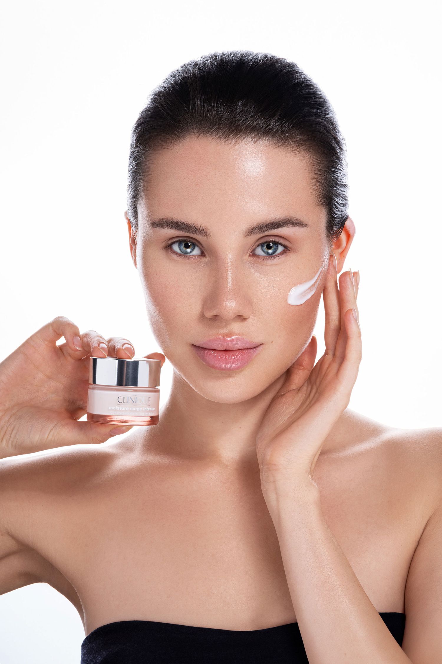 Beauty model applying moisturizing face cream while holding jar of skincare product against white background.