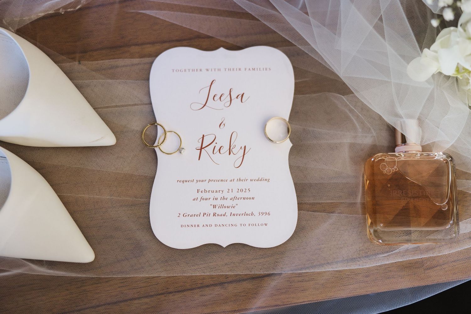 Elegant wedding invitation with rose gold lettering displayed alongside white heels and perfume bottle on wooden surface.