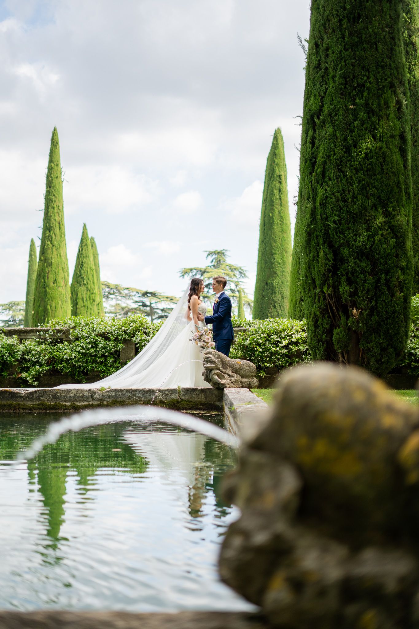 A couple stands on a stone bridge surrounded by tall cypress trees and reflecting pool in an elegant garden setting.