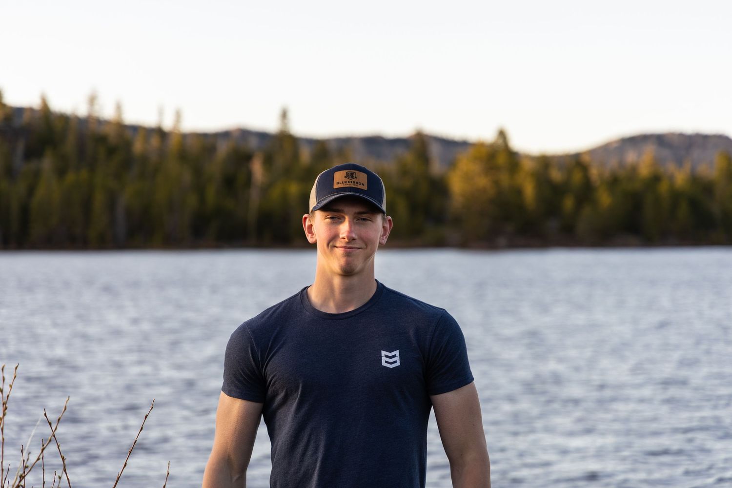 Young man in navy shirt and trucker hat smiling in front of a calm mountain lake at dusk.