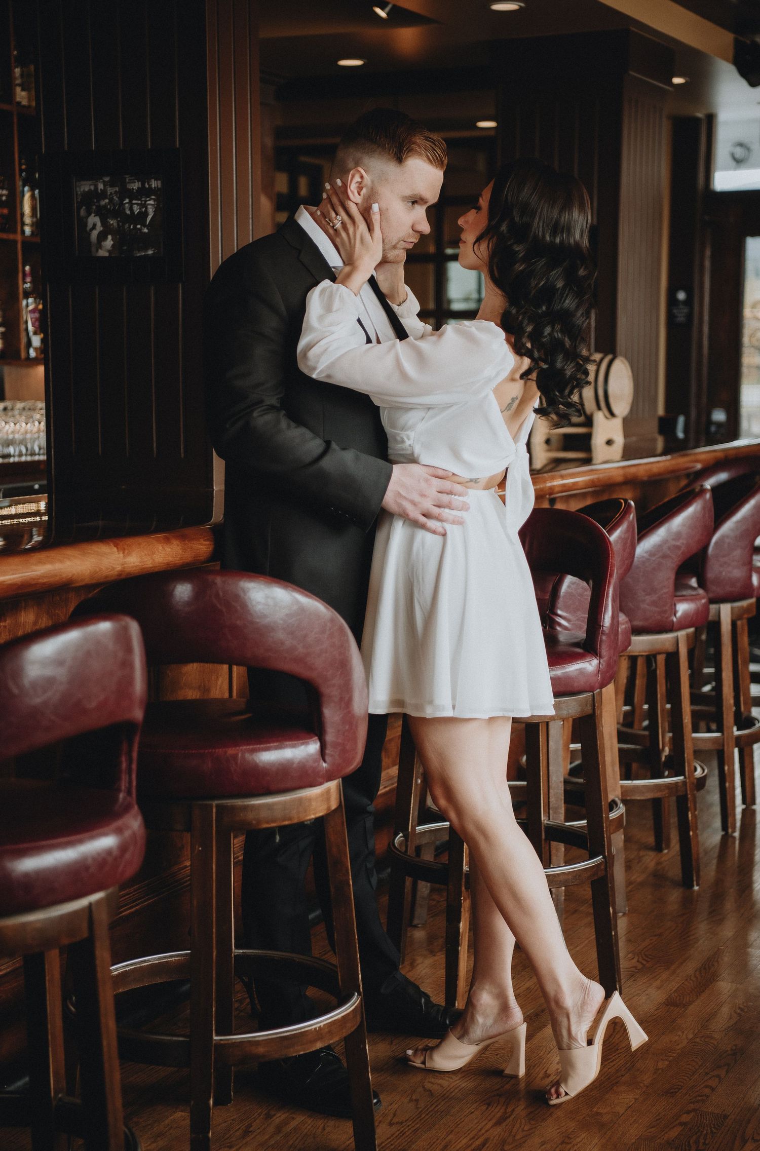 A romantic couple embraces at a vintage bar, she in a white dress and he in a dark suit.