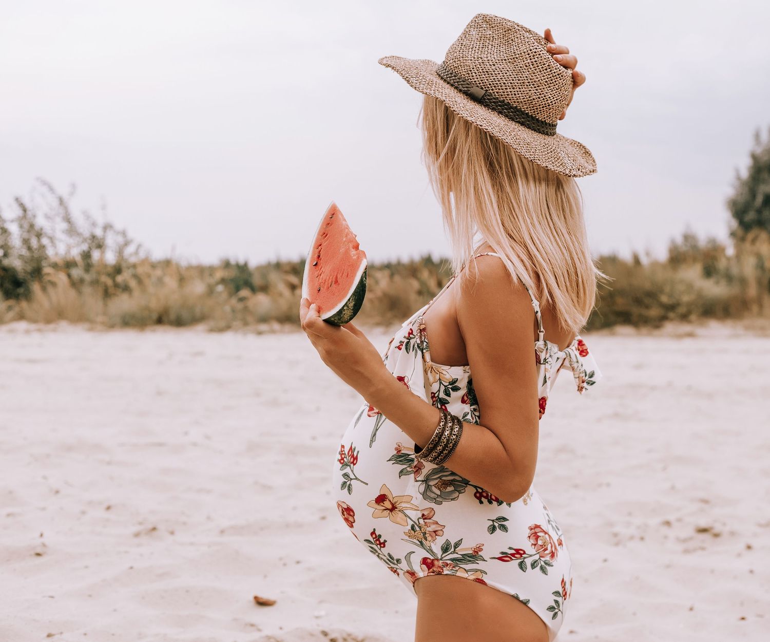 Woman in floral swimsuit and sun hat holds fresh watermelon slice on sandy beach during summer day.