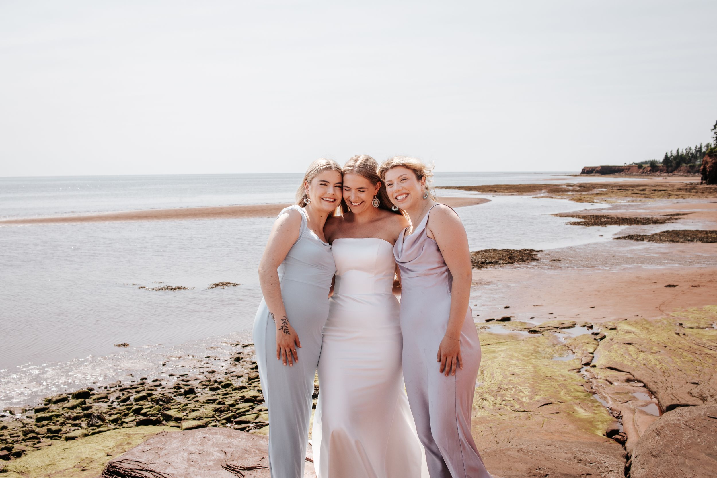 Three people wearing elegant white dresses pose together on a scenic beach at sunset with red cliffs in the background. Photo on PEI