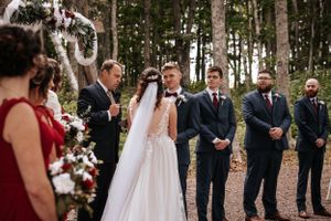 A wedding ceremony takes place in a wooded forest setting with a floral arch and wedding party in formal attire. Photo at Clinton Hills