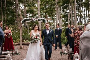 A joyful couple walks down an outdoor forest aisle decorated with white floral arch as guests look on. Photo at Clinton Hills