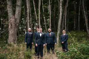 A group of men in formal navy suits stand together in a forest clearing surrounded by tall trees. Photo at Clinton Hills
