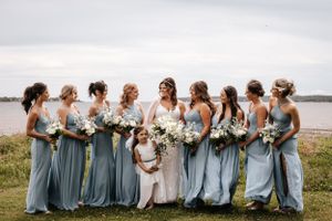 A bridal party in dusty blue bridesmaid dresses poses together by a serene waterfront on a cloudy day. Photo at Charlottetown PEI