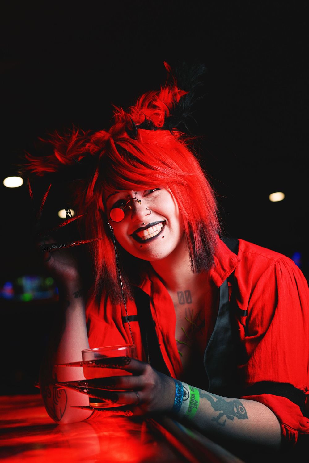 Person in red wig and clown makeup holding a drink under red lighting at a dark venue.