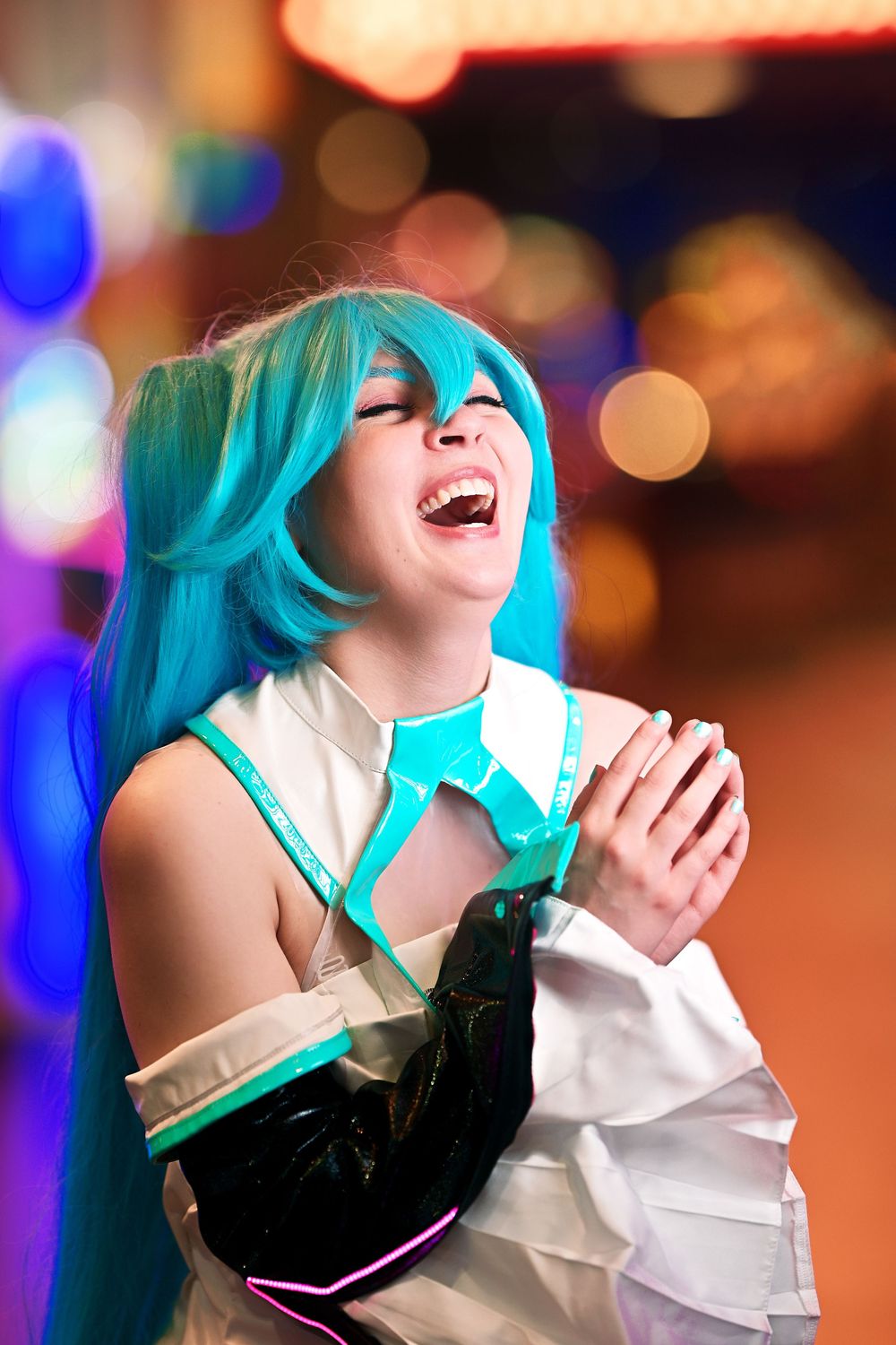 Young woman cosplaying as Hatsune Miku with teal twin-tail wig, laughing joyfully against a colorful bokeh background.