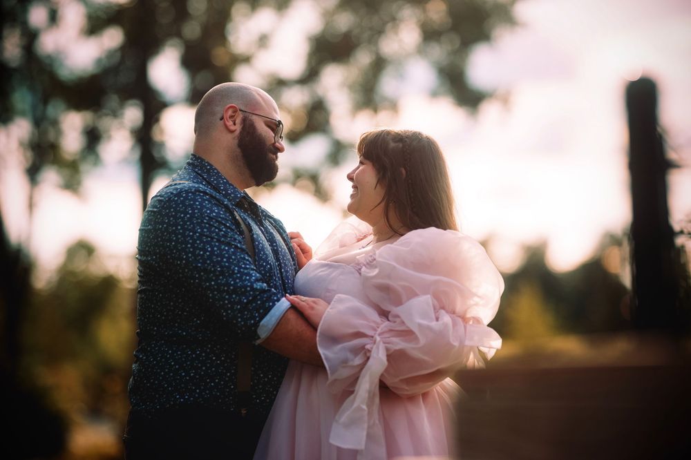 Couple embracing outdoors at sunset, man in blue jacket and woman in pink tulle dress, sharing a romantic moment.