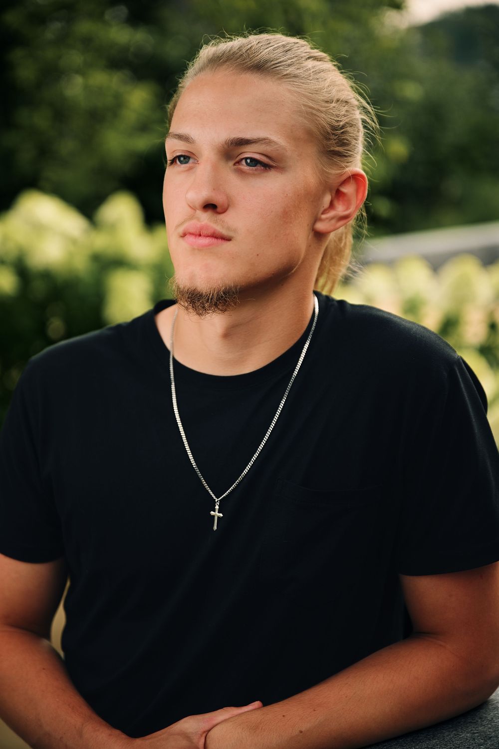 Young blonde man with a cross necklace wearing a black t-shirt, posing outdoors with green foliage background.