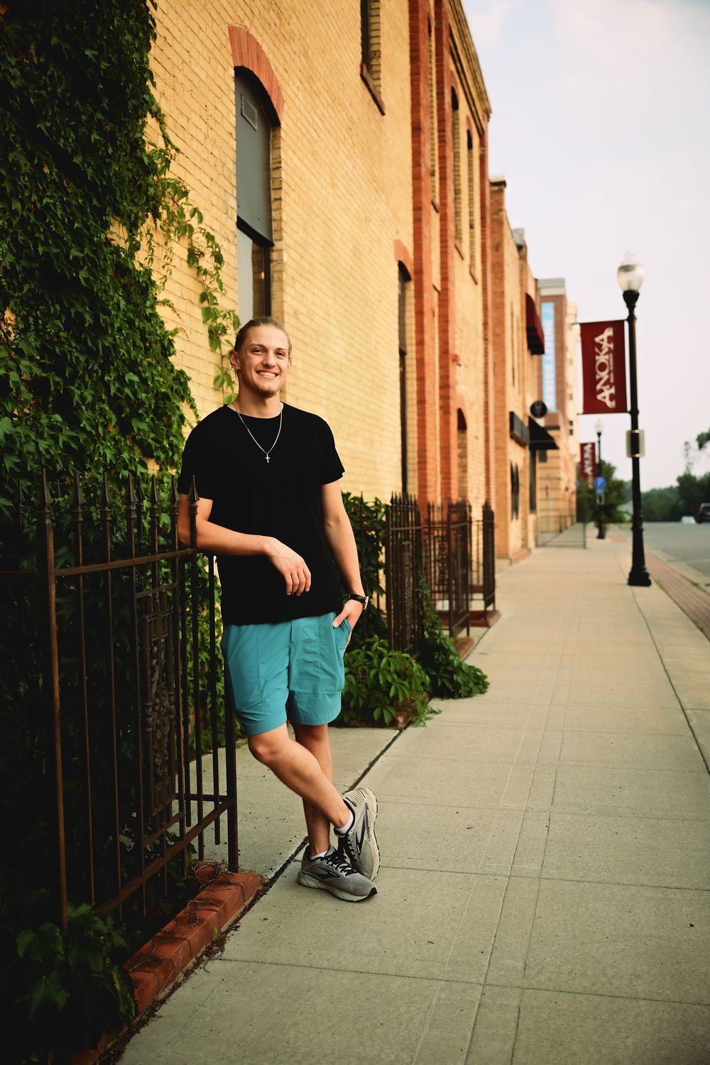 Smiling young man in black tee and teal shorts leaning on iron fence by brick building on urban sidewalk.