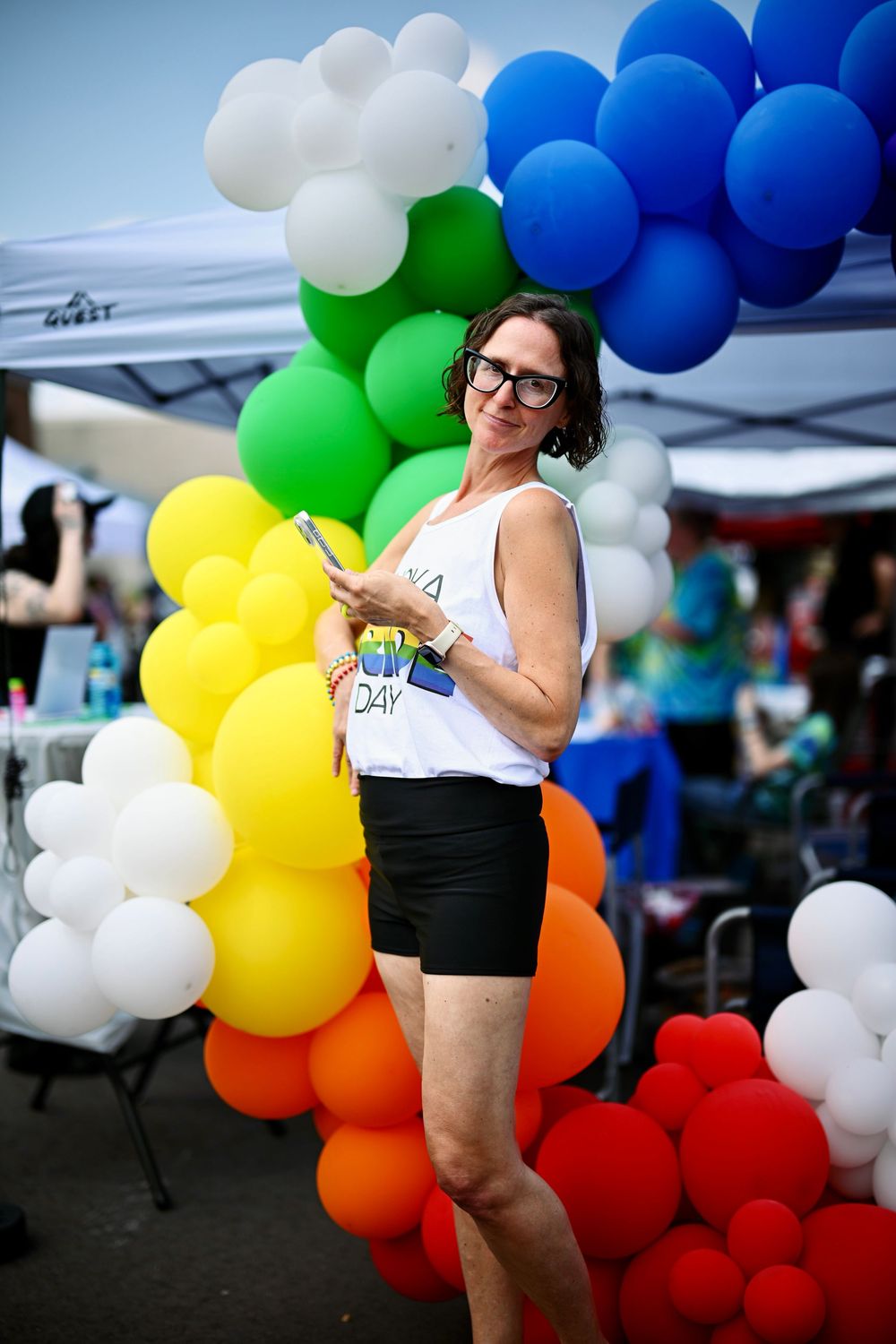 Woman in glasses poses beside colorful rainbow balloon arch at outdoor Pride festival event.
