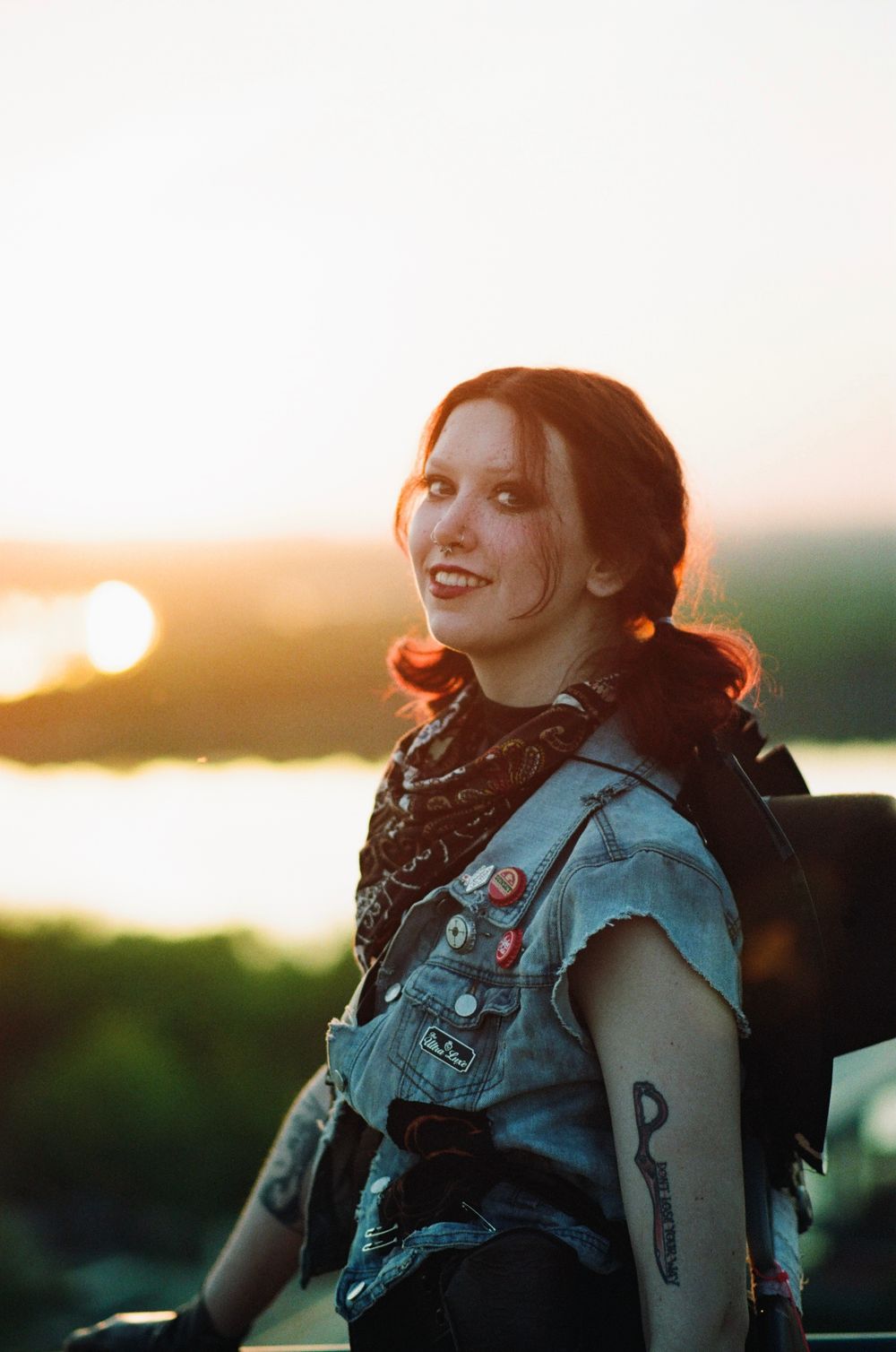 Young woman with red hair, denim vest, patches, and tattoos smiling at sunset outdoors.